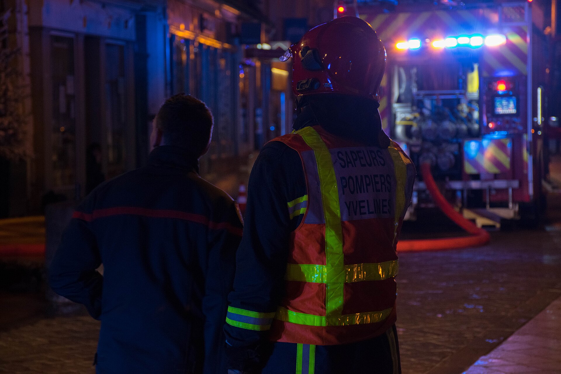 Deux sapeurs-pompiers de dos face au fourgon-pompe, gyrophares allumés